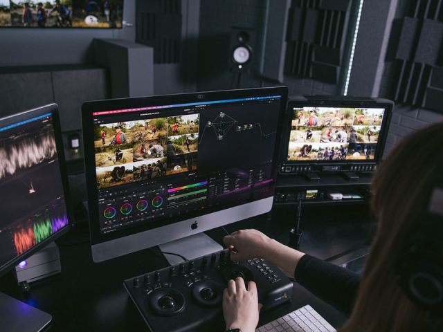 Post production editor sits at desk with 3 monitors editing with a forth large screen on the wall in background.